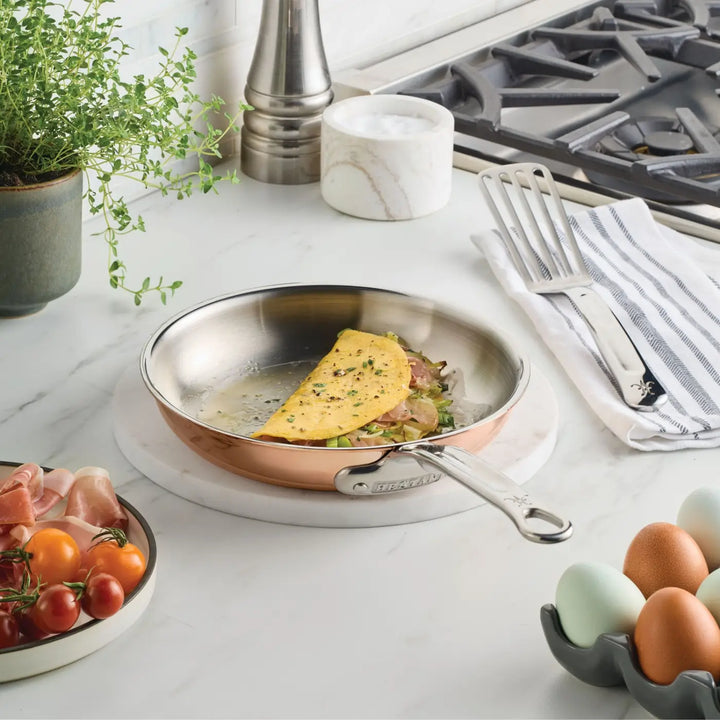 A perfectly half-folded omelet with herbs and filling sits in the CopperBond® Aura™ Polished Copper Skillet, its sleek stainless steel interior gleaming atop a marble counter surrounded by tomatoes, eggs, fresh herbs, a spatula, and a striped towel. Professional-grade cookware like the CopperBond® Aura™ Polished Copper Skillets helps you achieve delicious results every time—just as seen here with the gas stove visible in the background.