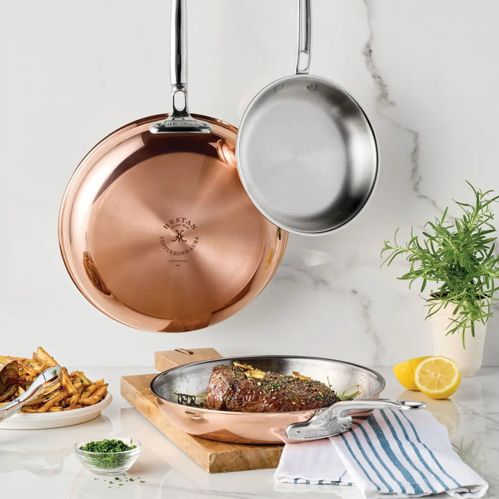 Two CopperBond® Aura™ Polished Copper Skillets, both professional-grade cookware, hang on the wall. On the counter below, a steak sizzles in another CopperBond® Aura™ skillet with a stainless steel interior, surrounded by fresh herbs, fries, a lemon, a striped towel, and various kitchen utensils.