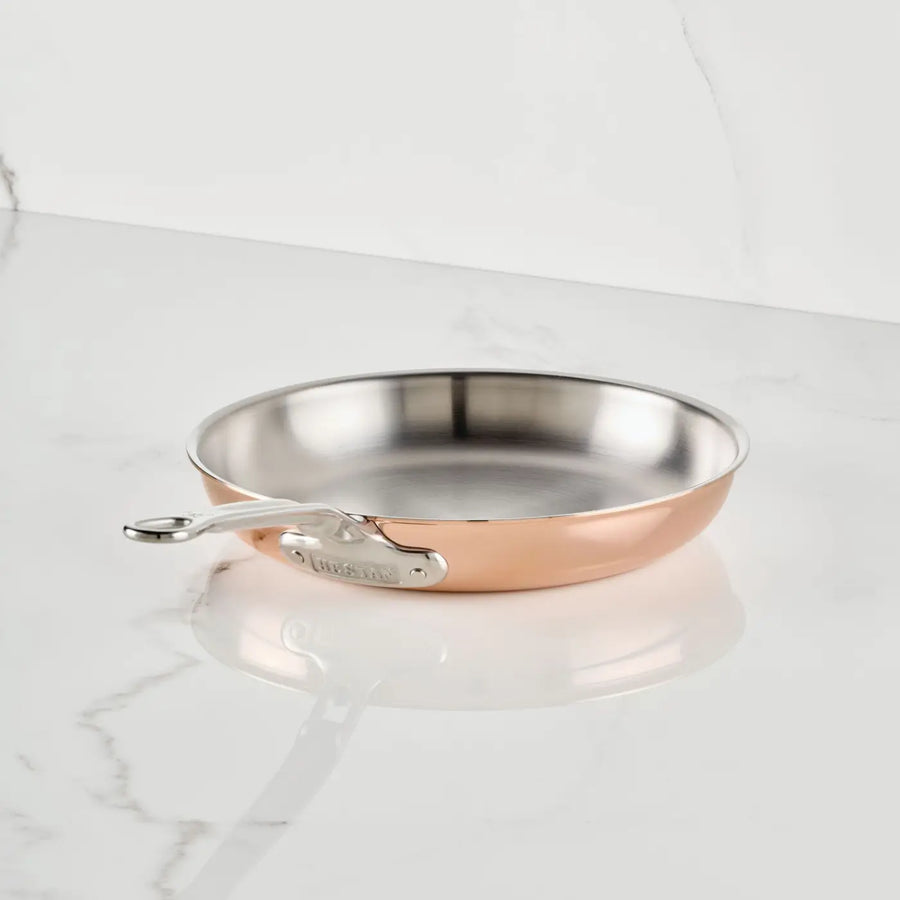 A CopperBond® Aura™ Polished Copper Skillet, featuring a stainless steel interior and metal handle, rests on a white marble countertop with delicate grey veining.