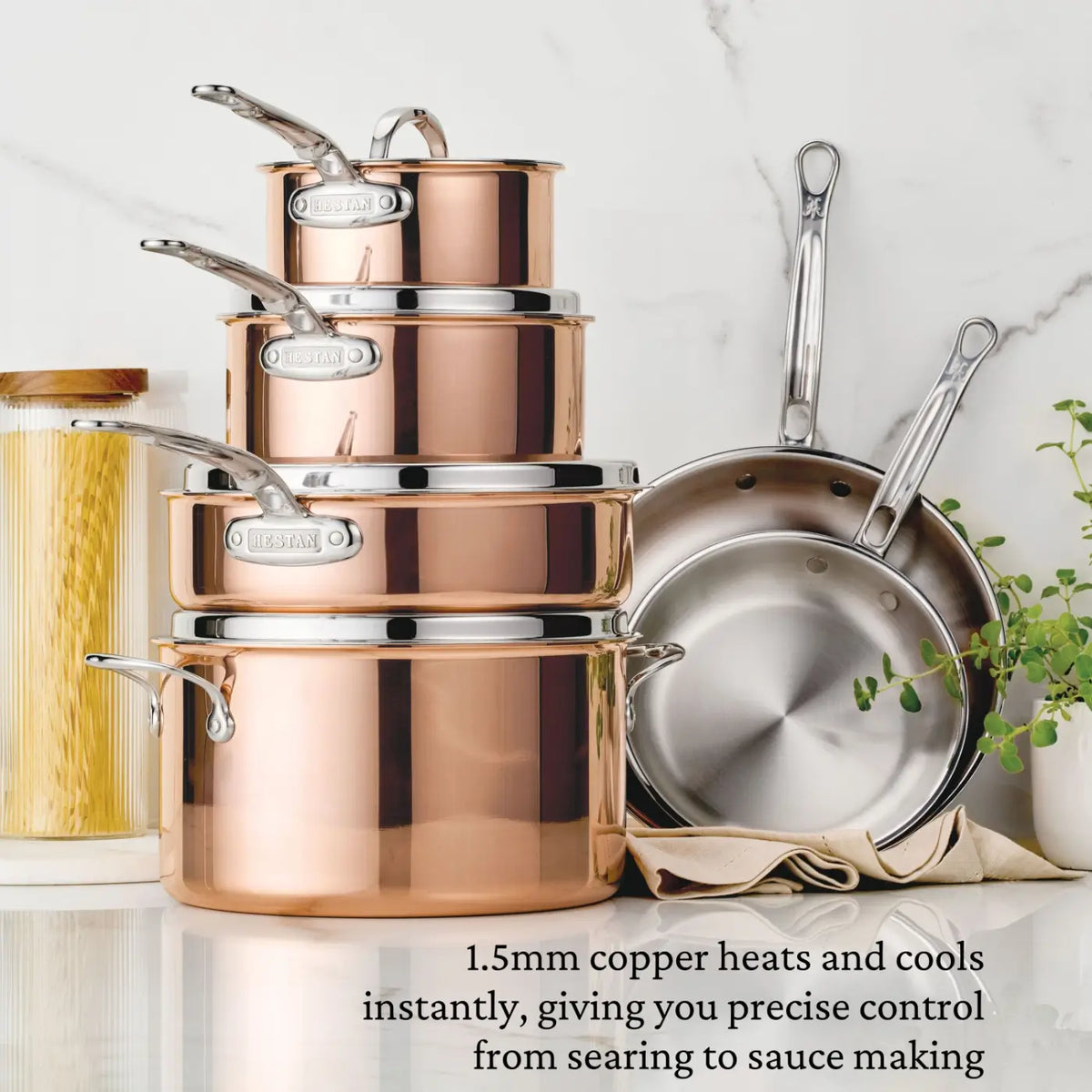 A collection of CopperBond® Aura™ Polished Copper Skillets, saucepans, a stockpot, and two frying pans is stacked neatly on a kitchen counter beside a jar of spaghetti and a small plant. Text highlights the cookware’s 1.5mm copper construction paired with a stainless steel interior.