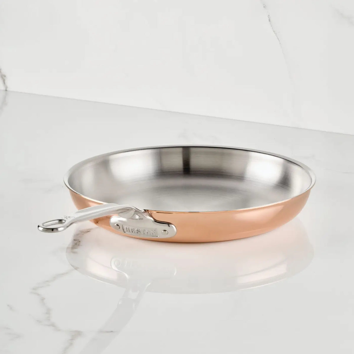 A Polished Copper Skillet from CopperBond® Aura™, featuring a stainless steel interior and a robust metal handle, rests on a white marble countertop with delicate gray veining, showcasing the enduring sophistication of CopperBond® Aura™ cookware.