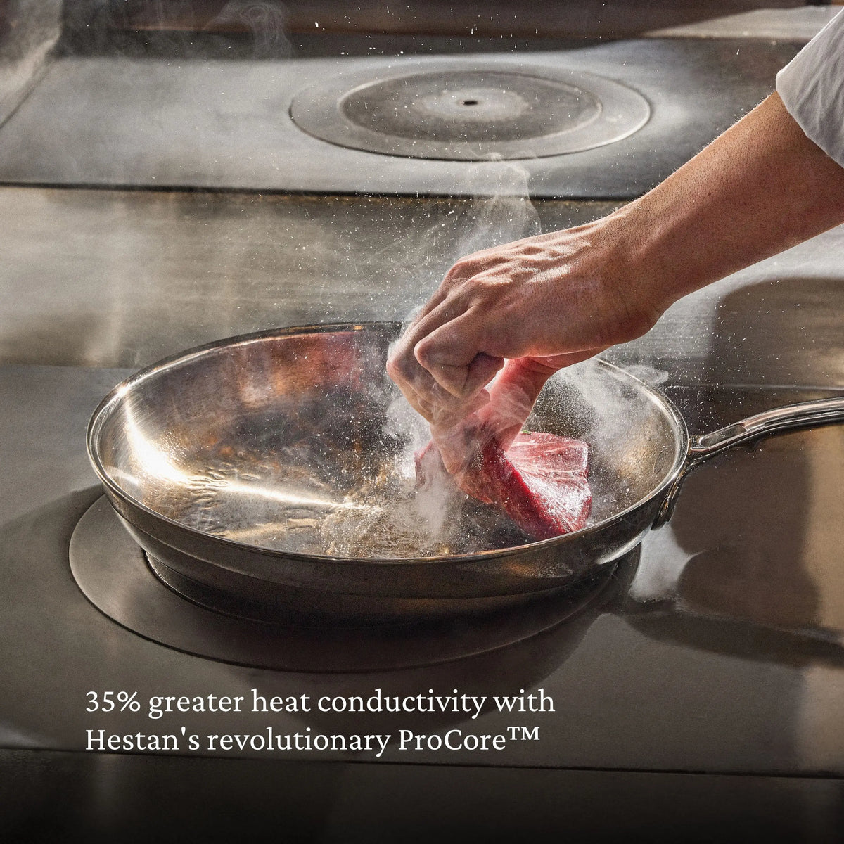 A hand sears raw meat in a stainless steel pan as steam rises. Text reads: “ProBond® Luxe™ Polished Clad Stainless Steel Signature Set, 7-Piece—35% greater heat conductivity with revolutionary ProCore™ tri-ply clad cookware.”.