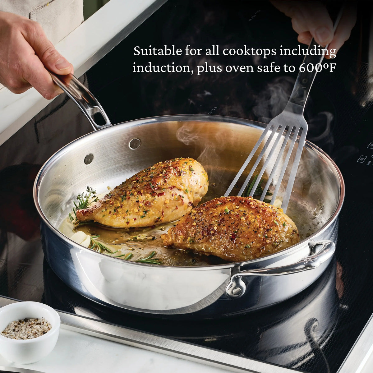 A cook uses a metal spatula to prepare two seasoned chicken breasts in the ProBond® Luxe™ Polished Clad Stainless Steel Signature Set, 5-Piece on an induction cooktop. Text: Suitable for all cooktops, including induction; oven safe to 600°F.