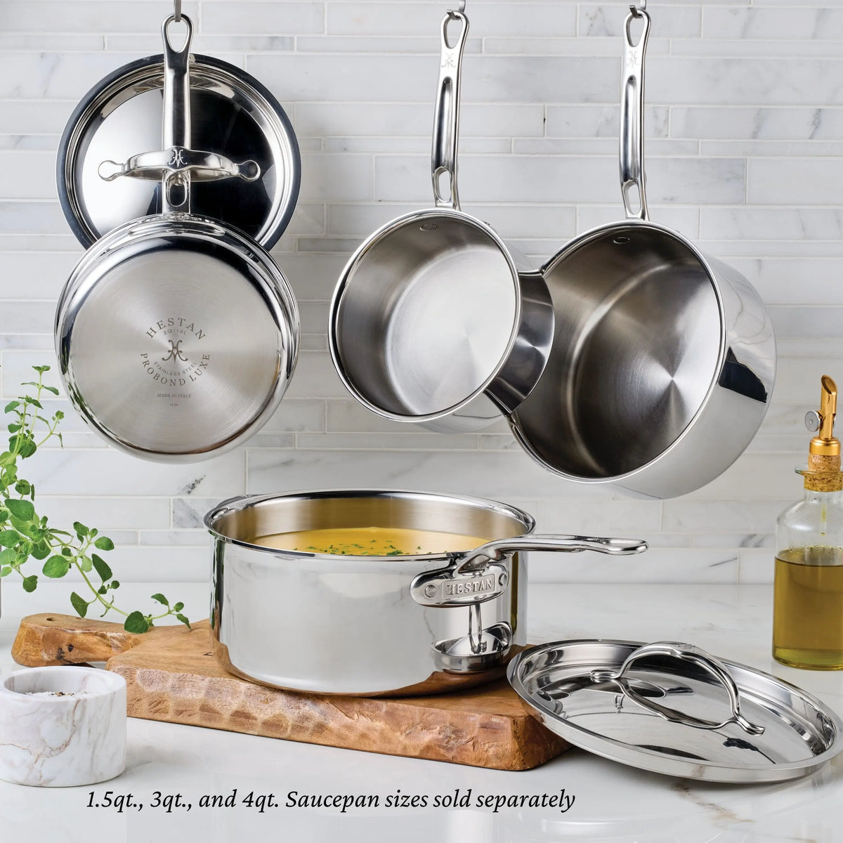 A ProBond® Luxe™ Polished Clad Stainless Steel Signature Set, 5-Piece is displayed with some saucepans and lids hanging on a wall rack and one filled with soup on a marble cutting board. A plant, oil bottle, and white dish sit nearby on the countertop.