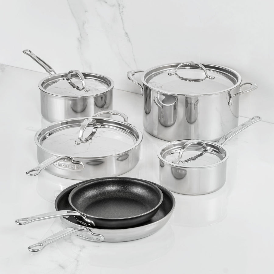 A ProBond Luxe® Polished Clad Stainless Steel 10-Piece Set, featuring four lidded pots and two black TITUM® nonstick frying pans, is elegantly arranged on a white marble countertop with a marbled white background—showcasing Italian craftsmanship at its finest.