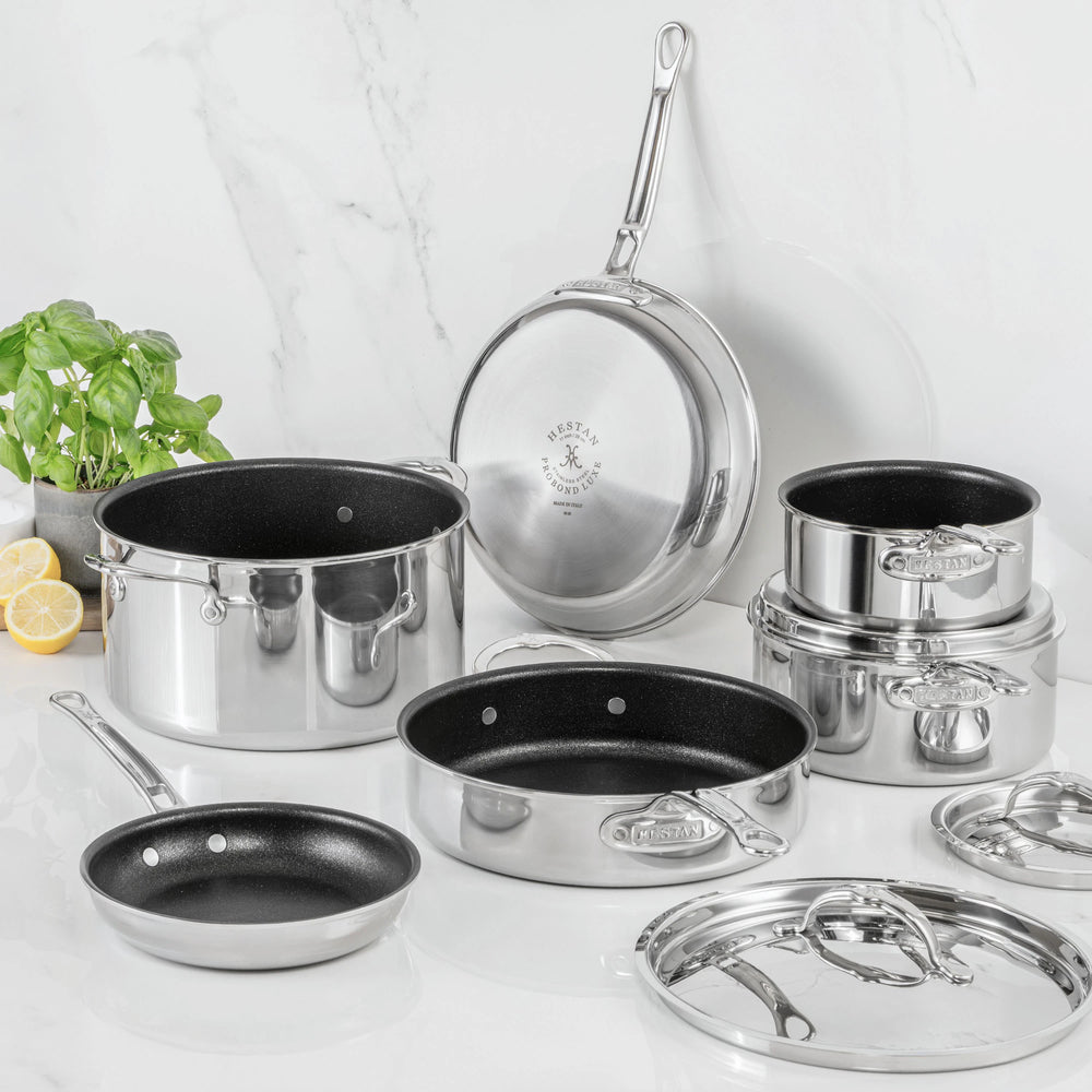 The ProBond Luxe® Polished Clad Stainless Steel 10-Piece Set, with TITUM® Nonstick, is displayed on a marble surface. Showcasing Italian craftsmanship, this cookware set features shiny stainless steel exteriors and black nonstick interiors. Various pots and pans—some with lids—are elegantly arranged, while a plant and sliced lemon in the background highlight the set’s sophisticated style and high performance.