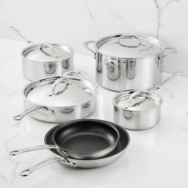 Five pieces of polished stainless steel cookware with lids, along with two nonstick frying pans from the ProBond Luxe® TITUM® Nonstick collection, are displayed on a white marble surface against a marble backdrop. Every piece in the Polished Clad Stainless Steel 10-Piece Set features sleek metal handles and a reflective finish.