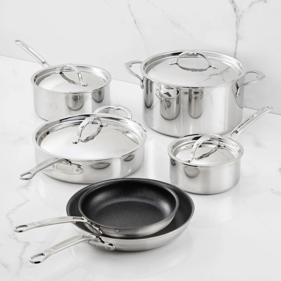 Five pieces of polished stainless steel cookware with lids, along with two nonstick frying pans from the ProBond Luxe® TITUM® Nonstick collection, are displayed on a white marble surface against a marble backdrop. Every piece in the Polished Clad Stainless Steel 10-Piece Set features sleek metal handles and a reflective finish.