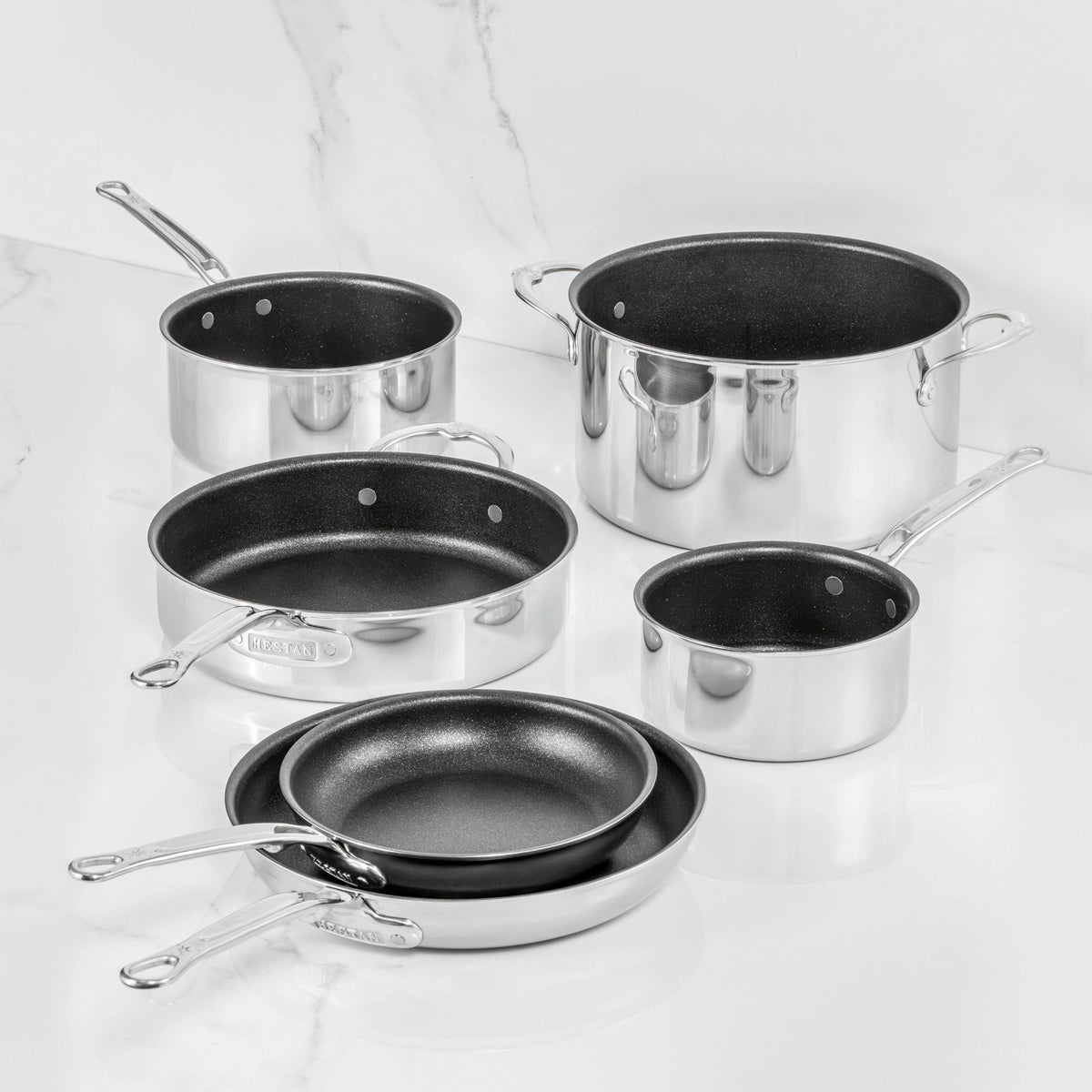 Inspired by Italian craftsmanship, the ProBond Luxe® Polished Clad Stainless Steel 10-Piece Set features TITUM® Nonstick interiors. This elegant collection includes two frying pans, two saucepans with handles, a sauté pan, and a large stock pot—all beautifully displayed on a white marble surface.