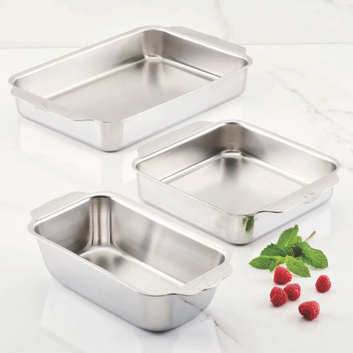An OvenBond Tri-Ply 3-Piece Baking Set by Hestan Provisions sits on a marble surface, surrounded by fresh raspberries and green mint leaves. This warp-resistant stainless steel set ensures durability while adding elegance to your kitchen.