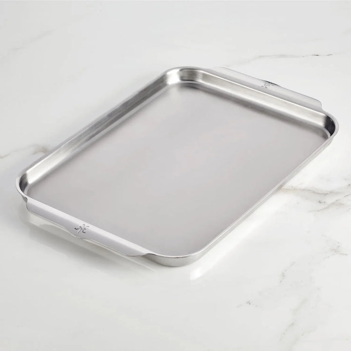 The Hestan Provisions OvenBond Tri-ply Half Sheet Pan, made from triple-bonded stainless steel, rests elegantly on a light marble surface. Its sleek rectangular design includes two side handles for easy handling.