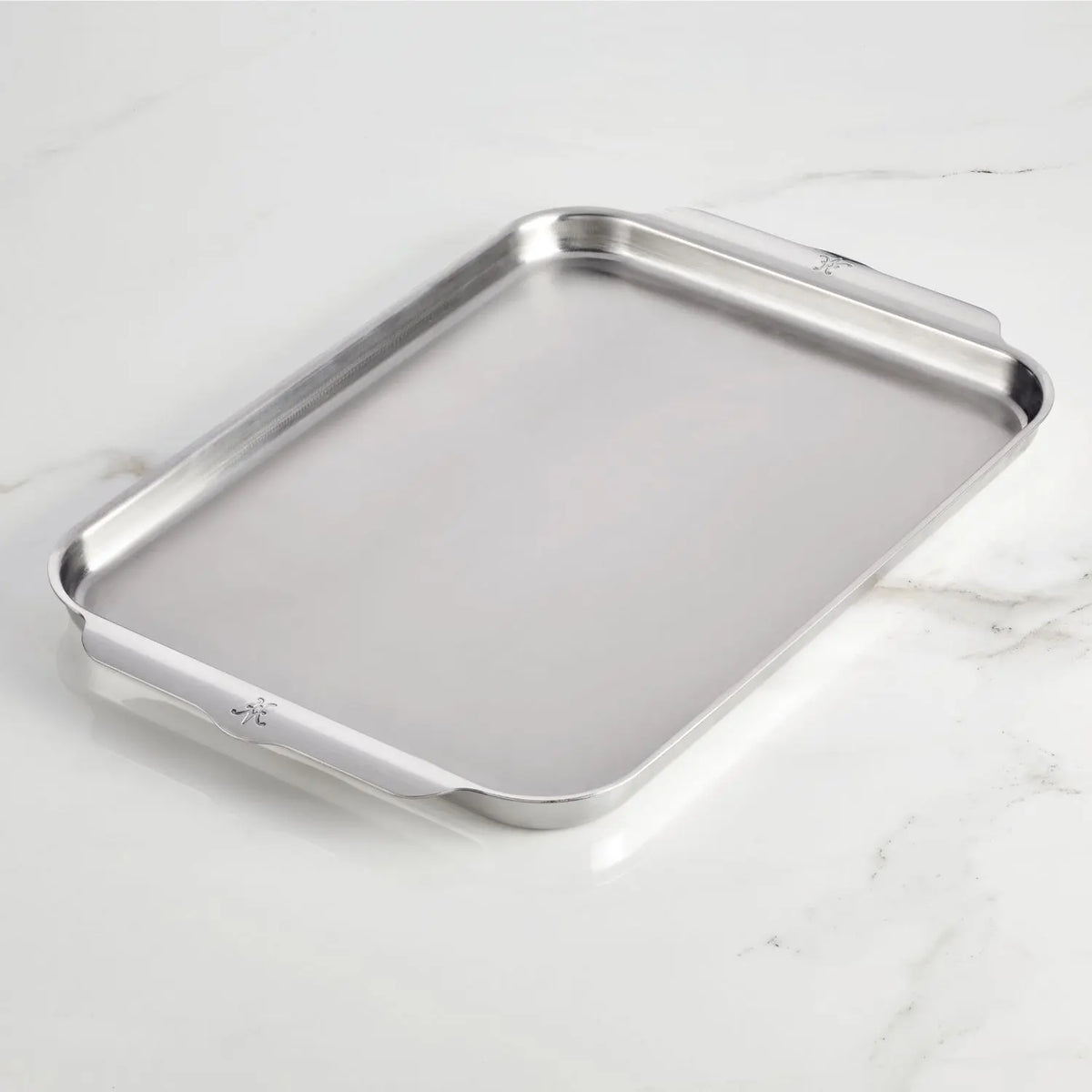 The Hestan Provisions OvenBond Tri-ply Half Sheet Pan, made from triple-bonded stainless steel, rests elegantly on a light marble surface. Its sleek rectangular design includes two side handles for easy handling.
