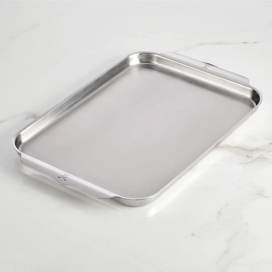 The Hestan OvenBond Tri-ply Half Sheet Pan & Rack (13"x18") with raised edges and two flat handles rests on a white marble surface.