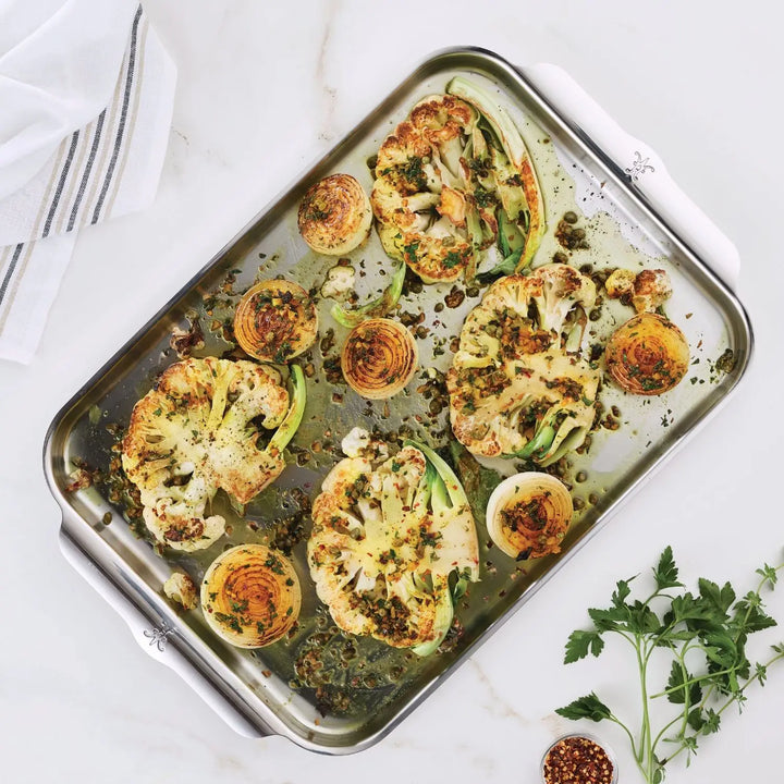 The Hestan Provisions OvenBond Tri-ply Half Sheet Pan evenly roasts vegetables like cauliflower and halved Brussels sprouts on a marble surface, garnished with herbs. Nearby, theres a striped cloth and a parsley sprig.