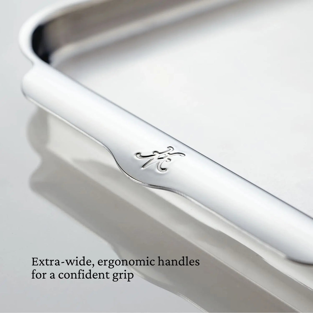 Close-up of the OvenBond Tri-ply Half Sheet Pan from Hestan Provisions, crafted from triple-bonded clad stainless steel with an engraved H and a curved edge. A text overlay reads, Extra-wide, ergonomic handles for a confident grip, against a soft, elegant background.