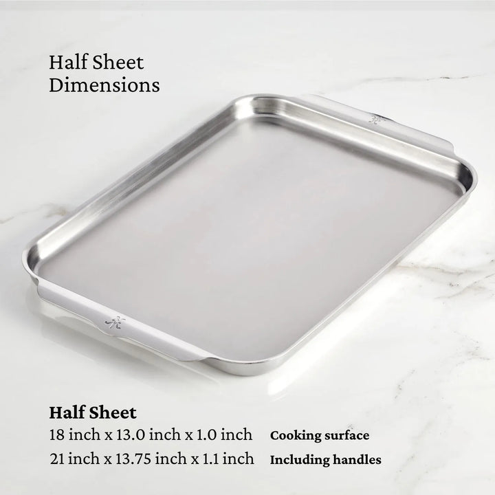 A stainless steel OvenBond Tri-ply Half Sheet Pan by Hestan Provisions sits on a marble surface. Text reads Half Sheet Dimensions with 18 x 13 x 1 inches for the cooking area, and 21 x 13.75 x 1.1 inches including handles, crafted with triple-bonded clad stainless steel.