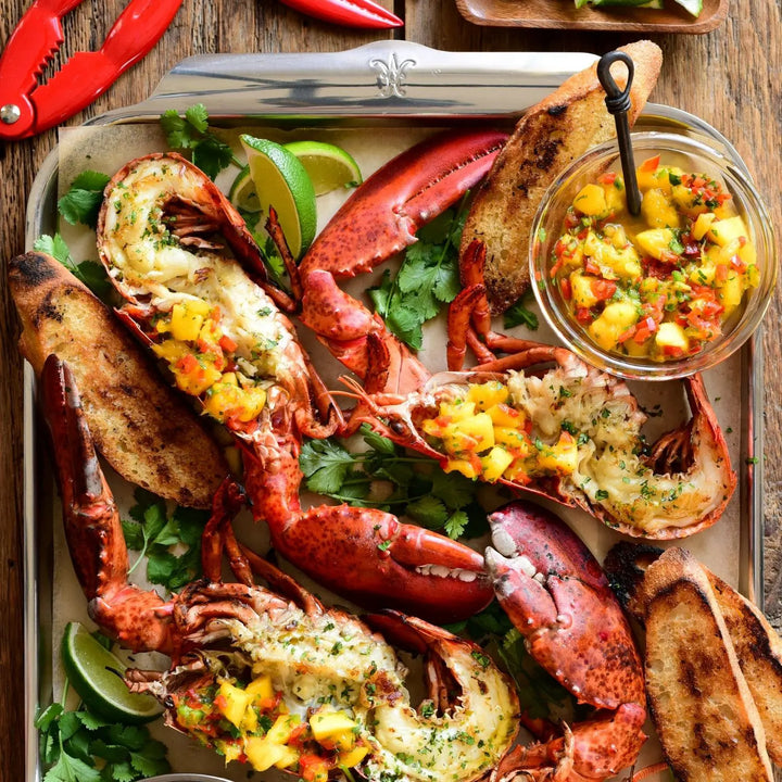 Grilled lobster halves with mango salsa are served on toasted bread with lime wedges and cilantro, beautifully arranged on the Hestan Provisions OvenBond Tri-ply Half Sheet Pan. Red lobster claw crackers and a small bowl of salsa with a spoon adorn the background.