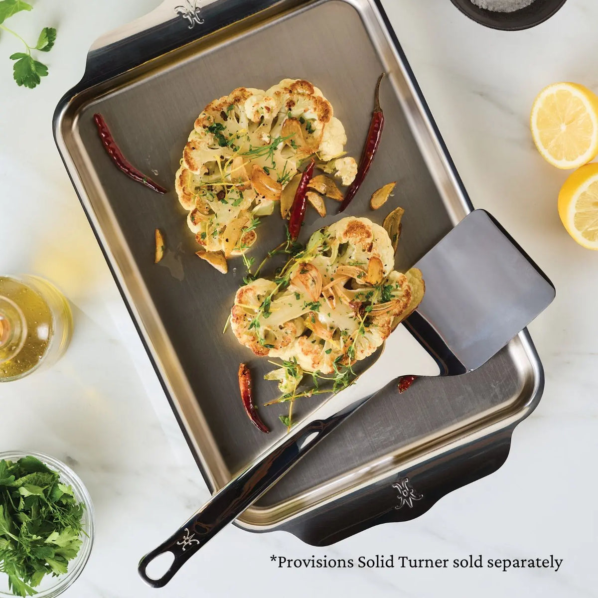 The Hestan Provisions OvenBond Tri-ply Quarter Sheet Pan holds roasted cauliflower steaks garnished with herbs and garlic. Red chilies are scattered on the pan, which is oven safe. A spatula lies beside it with half a lemon, a glass of oil, and fresh herbs nearby.