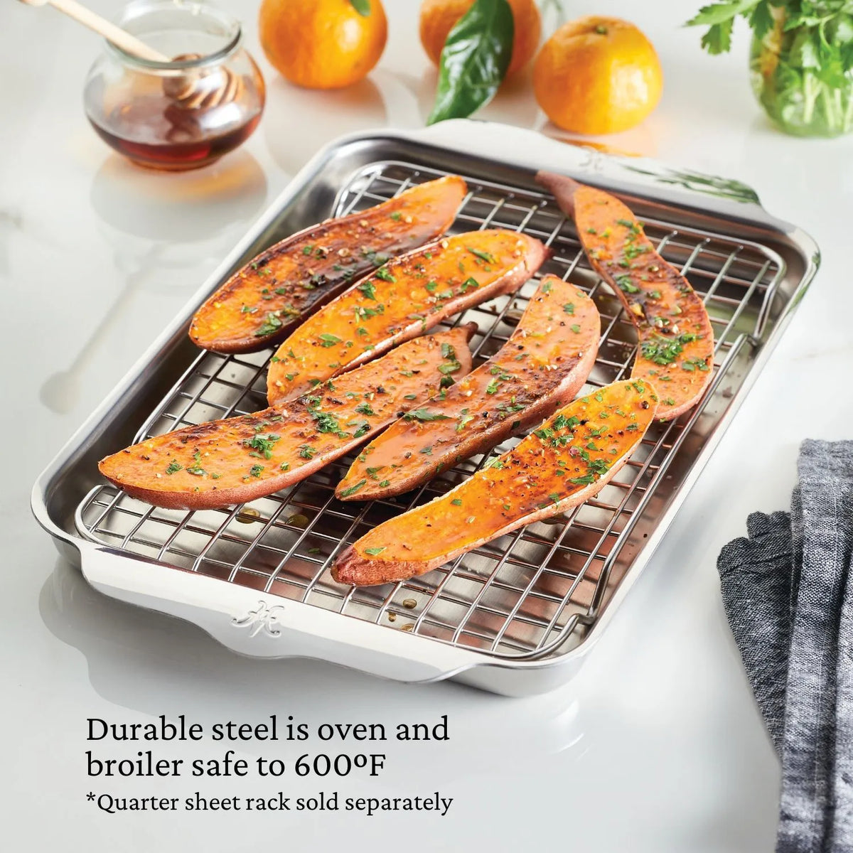 The OvenBond Tri-ply Quarter Sheet Pan by Hestan Provisions holds roasted sweet potato wedges on a wire rack. Its placed on a white surface, surrounded by scattered parsley and oranges. The text reads, Stainless steel is oven safe to 600°F.