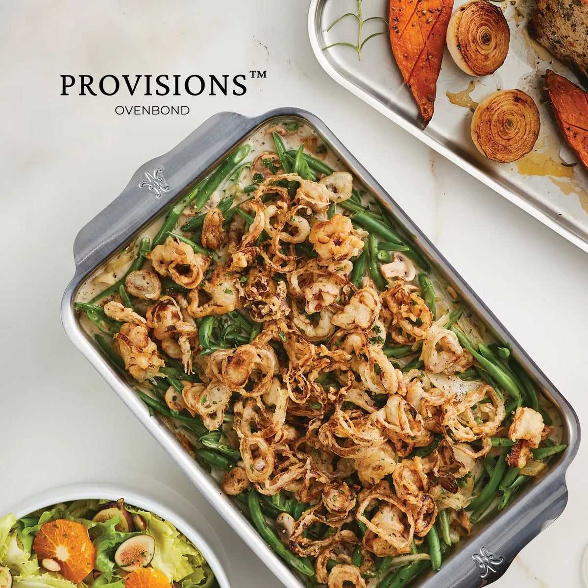 A Hestan Provisions OvenBond Tri-ply Quarter Sheet Pan filled with green bean casserole and topped with crispy fried onions is on a table, accompanied by a platter of roasted vegetables and a citrus green salad.