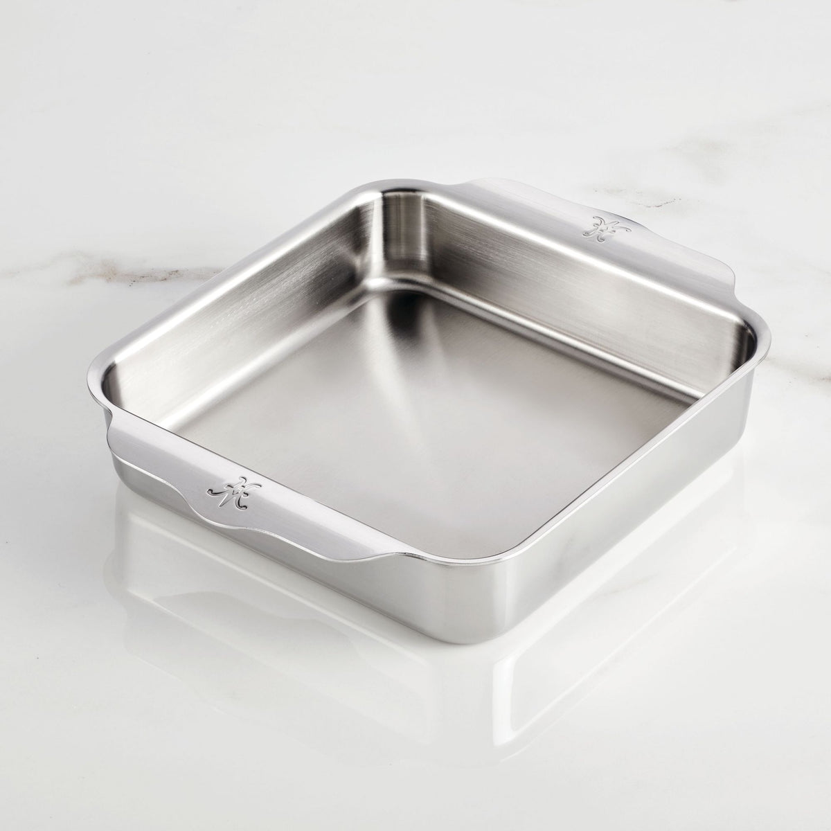 The Hestan Provisions OvenBond Tri-ply Square Baker, crafted with stainless steel and featuring two extended handles, rests on a smooth white marble surface. It boasts a polished reflective finish and is oven safe.