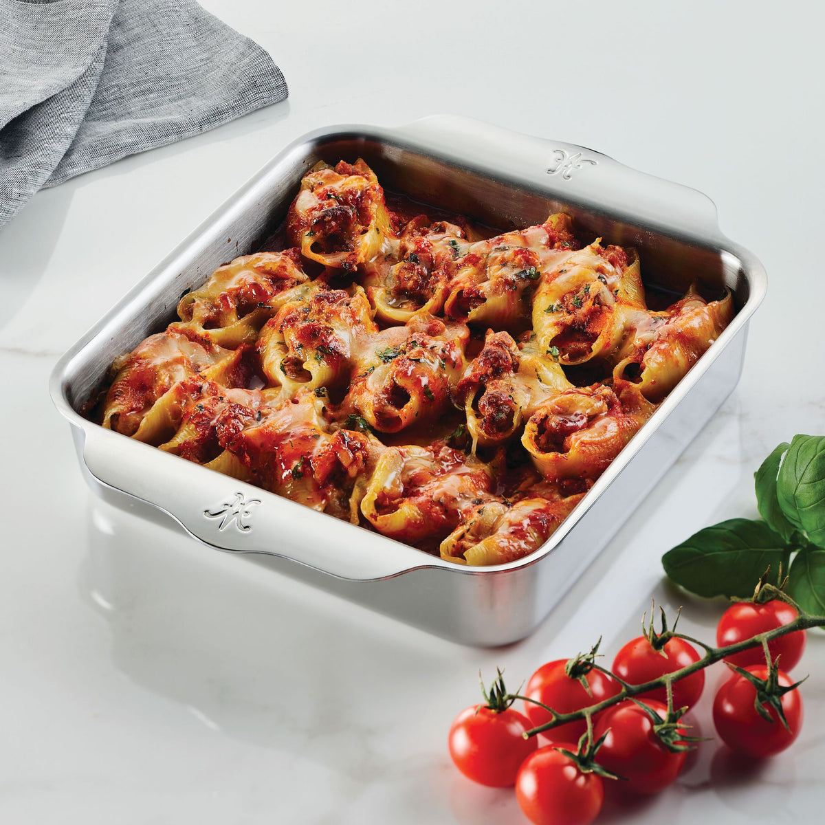 Baked stuffed pasta shells with melted cheese and sauce fill a metal dish from the OvenBond Tri-Ply 3-Piece Baking Set by Hestan Provisions. Cherry tomatoes and basil leaves complement it on a white marble surface, alongside a gray napkin. Warp-resistant elegance for every kitchen.