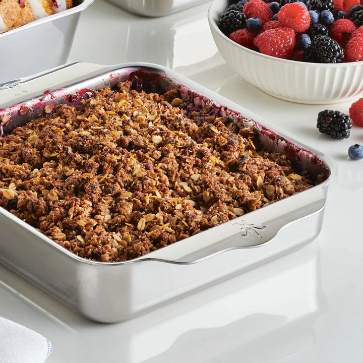 The Hestan Provisions OvenBond Tri-ply Square Baker presents a baked crumble with a golden-brown topping. Beside it, theres a white bowl filled with raspberries, blueberries, and blackberries. Other dishes are partially seen in the background on a white surface.