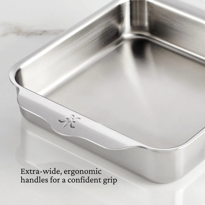 The image showcases the Hestan Provisions OvenBond Tri-ply Square Baker. Its stainless steel design features extra-wide, ergonomic handles with logo engraving for a confident grip. The text reads: Extra-wide, ergonomic handles for a confident grip, set against a white marble background.