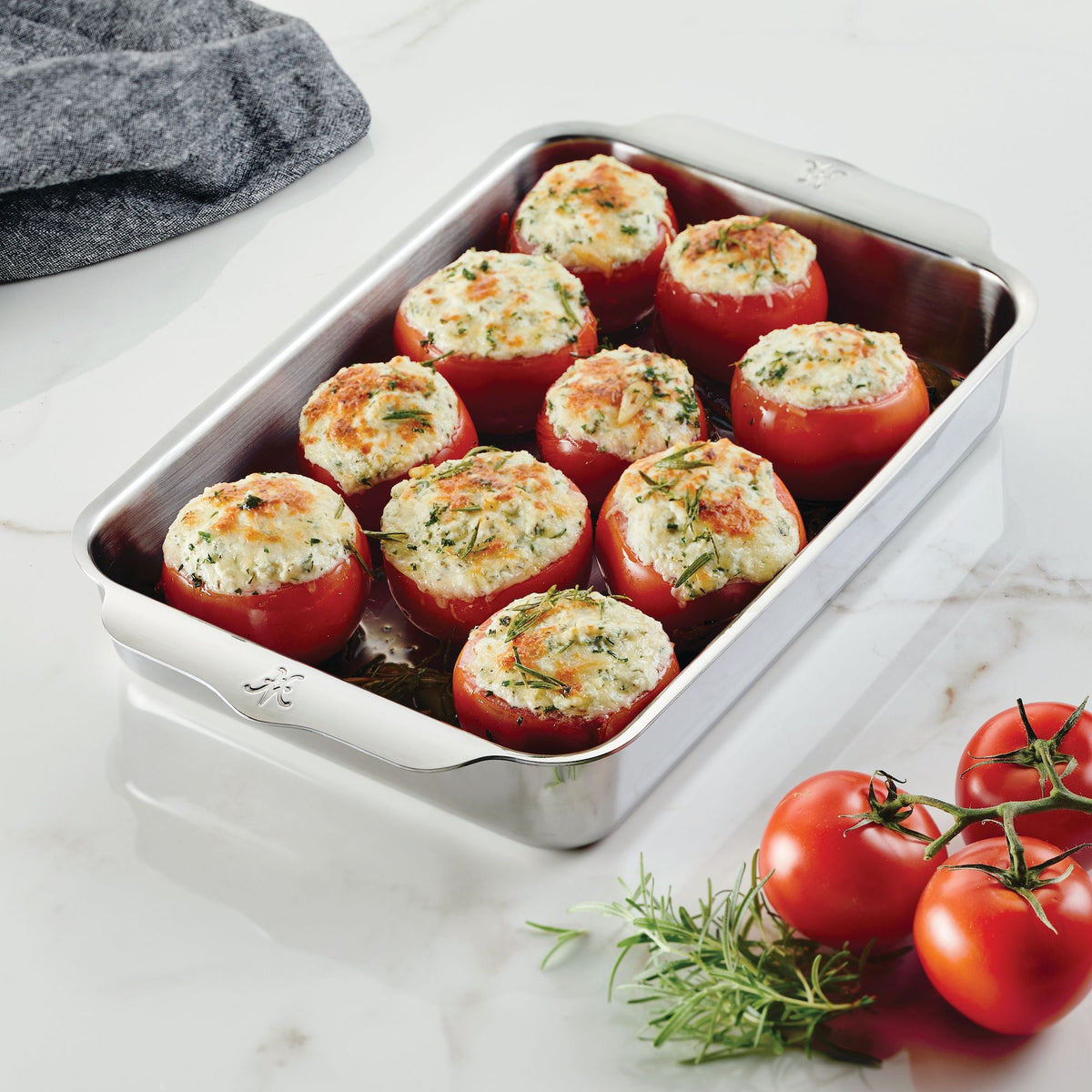 A metal baking dish from Hestan Provisions OvenBond Tri-Ply 3-Piece Baking Set holds stuffed tomatoes in a white mixture, topped with herbs. The warp-resistant dish sits on marble, surrounded by fresh tomatoes and rosemary sprigs, with a dark cloth peeking out from the corner.