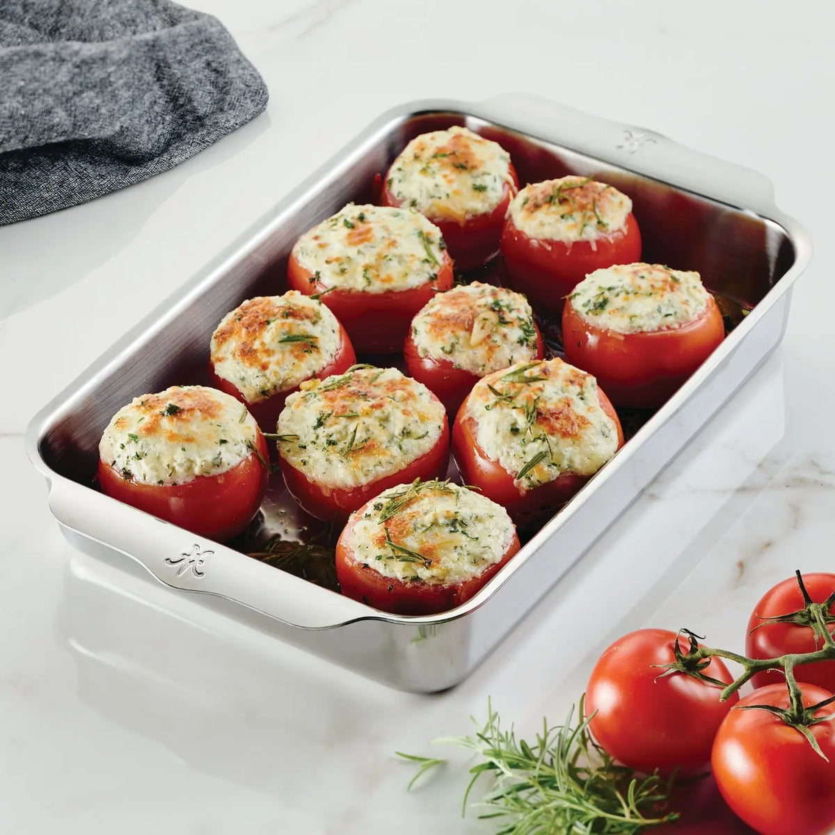 The Hestan Provisions OvenBond Tri-ply Rectangular Baker fits eight stuffed tomatoes topped with cheese and herbs. On a marble countertop, this dish is paired with a sprig of rosemary and four whole tomatoes.