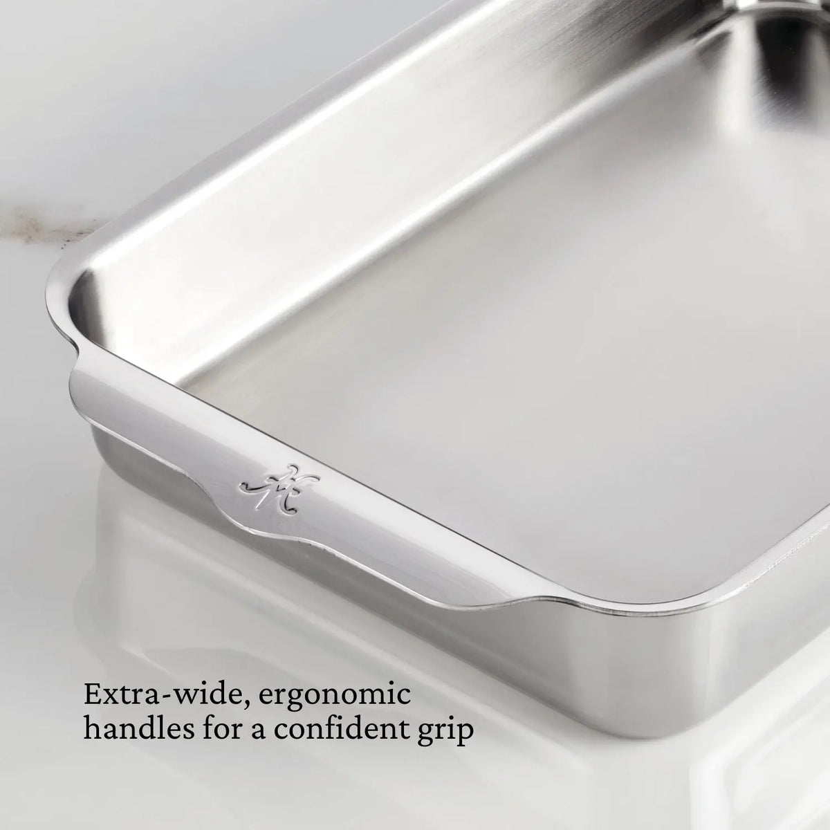 A close-up of the Hestan Provisions Host and Roast 8-piece set shows a stainless steel roasting pan with extra-wide, ergonomic handles for a confident grip. The durable bakeware rests on a white surface.
