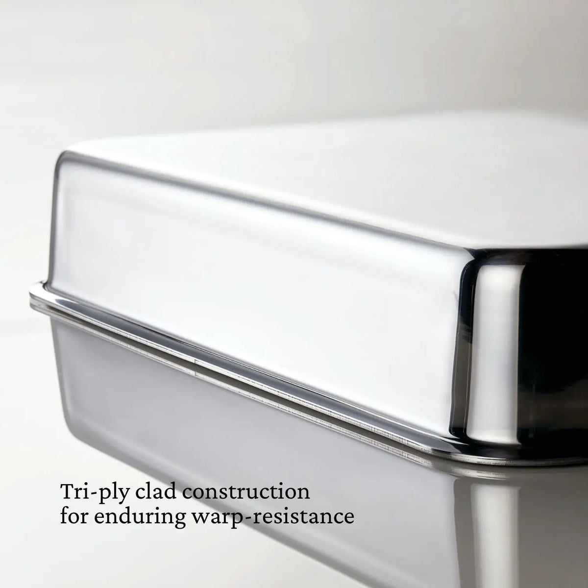 A close-up of the corner of a Hestan Provisions Host and Roast, 8-piece Set stainless steel pan with shiny, reflective surfaces. Text reads: Tri-ply clad construction for enduring warp-resistance—ideal for holiday prep.