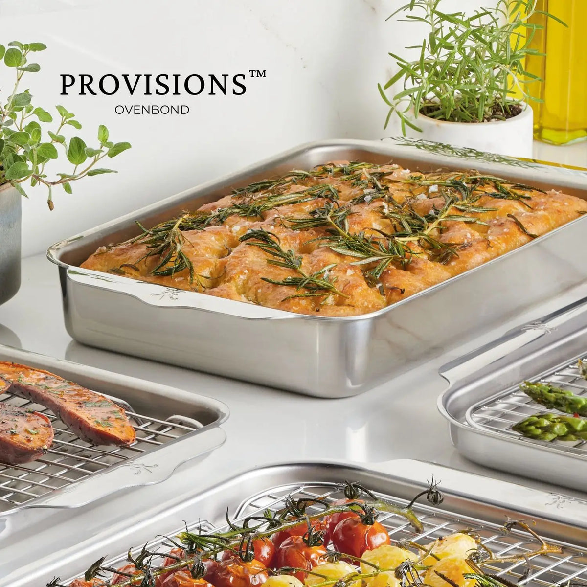A stainless steel OvenBond Tri-ply Rectangular Baker holds roasted chicken garnished with rosemary on a kitchen counter. Surrounding trays boast grilled asparagus, tomatoes, and sausages. Fresh herbs and oil complete the scene. The text reads Hestan Provisions.