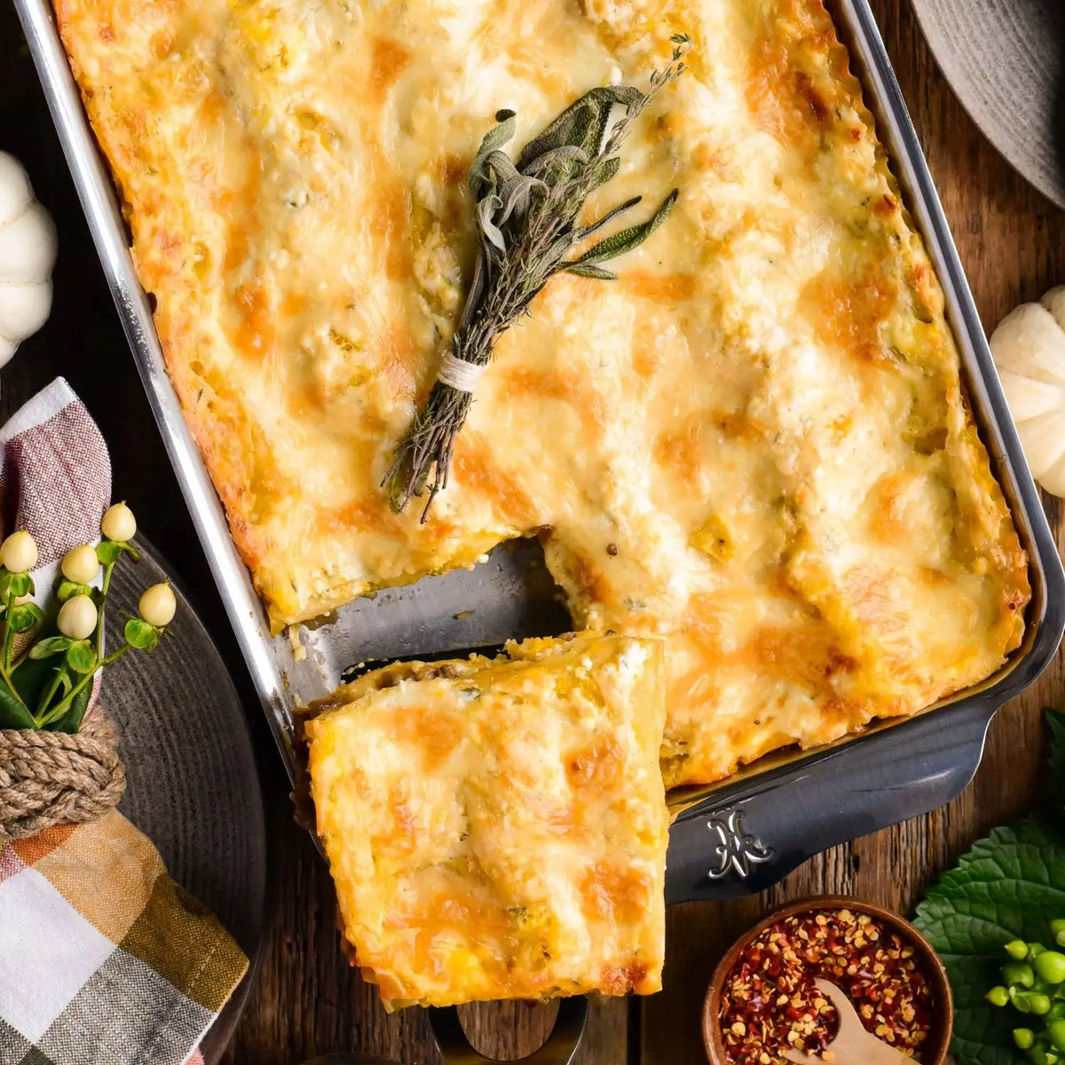 A baked lasagna in an OvenBond Tri-ply Rectangular Baker sits on a wooden table. A slice is partially removed, revealing layers. A sprig of herbs lies on top. Nearby are red chili flakes in a small bowl, a cloth napkin, and decorative items from Hestan Provisions.