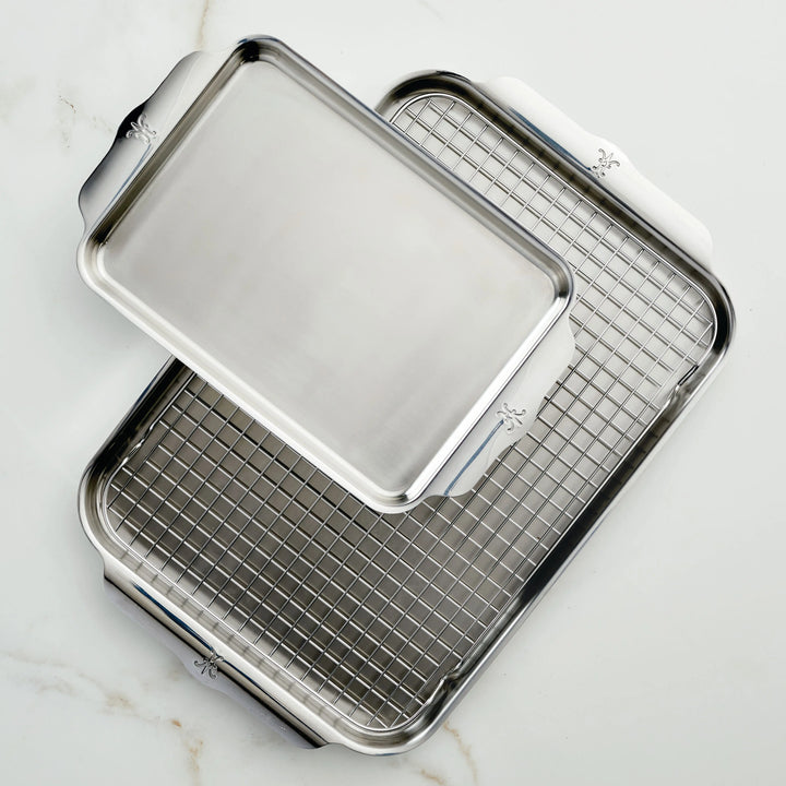 Two stainless steel sheet pans from the Hestan Provisions OvenBond Tri-ply 3-Piece Set are displayed on a white marble surface. One tray is stacked diagonally atop the other, which holds a metal cooling rack. Both trays have handles on the short sides.