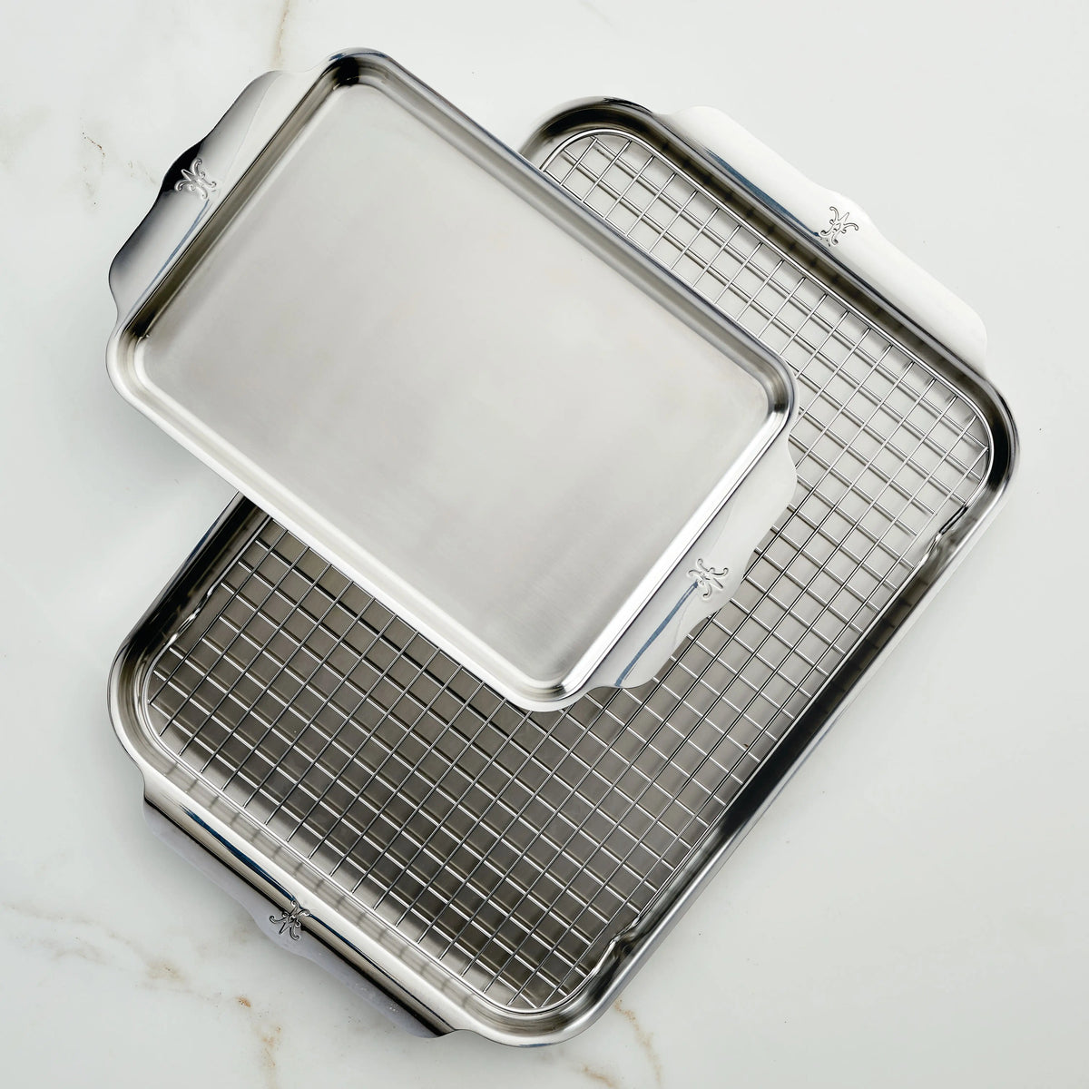 Two stainless steel sheet pans from the Hestan Provisions OvenBond Tri-ply 3-Piece Set are displayed on a white marble surface. One tray is stacked diagonally atop the other, which holds a metal cooling rack. Both trays have handles on the short sides.