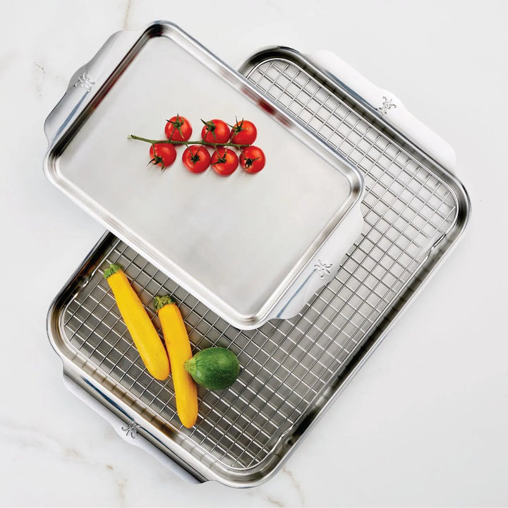 The OvenBond Tri-ply 3-Piece Set from Hestan Provisions features two stainless steel baking trays. One has a wire rack, and the other, with embossed handles, holds cherry tomatoes and zucchinis. Made with an aluminum core for warp resistance, these roasting pans sit on a white marble surface.