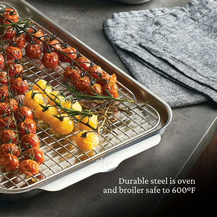 The Hestan Provisions OvenBond Tri-ply 3-Piece Set includes a warp-resistant roasting pan with a metal rack, perfect for holding red and yellow cherry tomatoes seasoned with herbs. Folded gray kitchen towels lie nearby. Durable steel is oven and broiler safe up to 600°F.