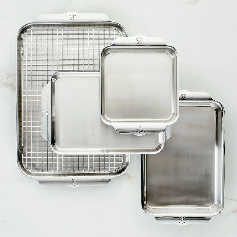 The Hestan Provisions OvenBond Tri-ply 5-Piece Set, featuring stainless steel bakeware pans in various sizes and a metal cooling rack, is shown with raised edges and decorative handles on a white surface.