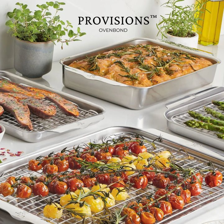 A kitchen scene highlights the Hestan Provisions OvenBond Tri-ply 5-Piece Set, showcasing roasted vegetables, bread topped with rosemary, and sausages. Cherry tomatoes and yellow peppers add color while potted plants and condiments enhance the background charm.