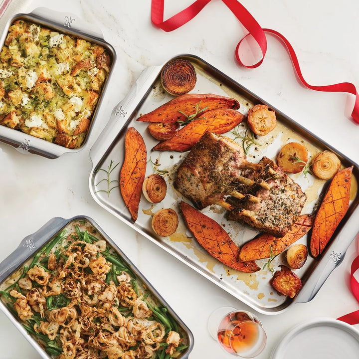 The table features a delightful meal: herb roasted meat, sweet potato wedges, and onions on an OvenBond Tri-ply tray from Hestan Provisions; a cheese-topped casserole; green beans with crispy onions, paired with pink liquid in a glass and adorned with a decorative red ribbon.