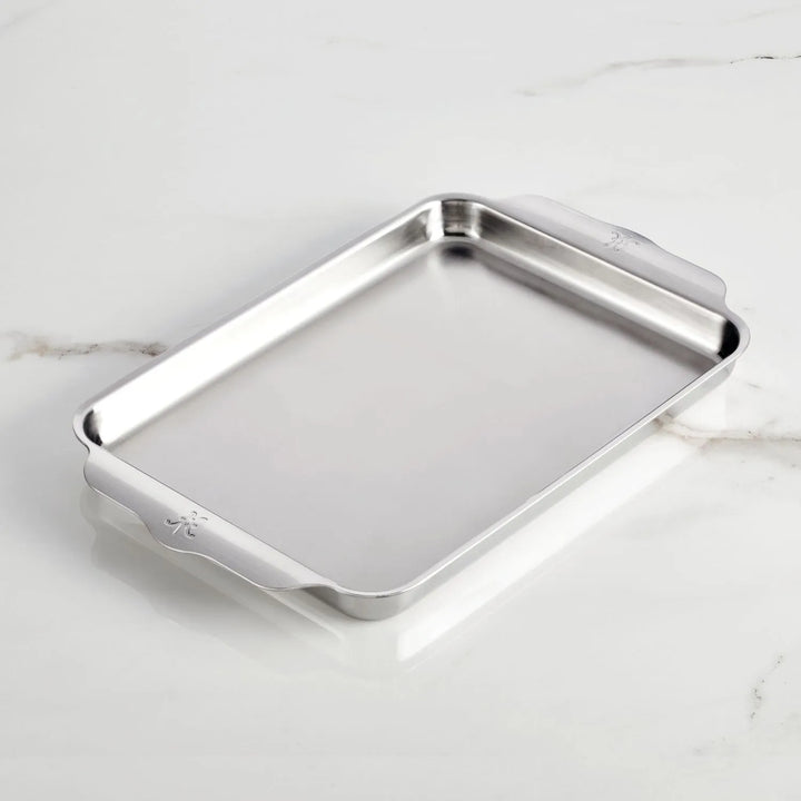 A Hestan OvenBond Tri-Ply Medium Sheet Pan & Rack, 12-in x 15-in, with raised edges and side handles rests on a white marble surface.