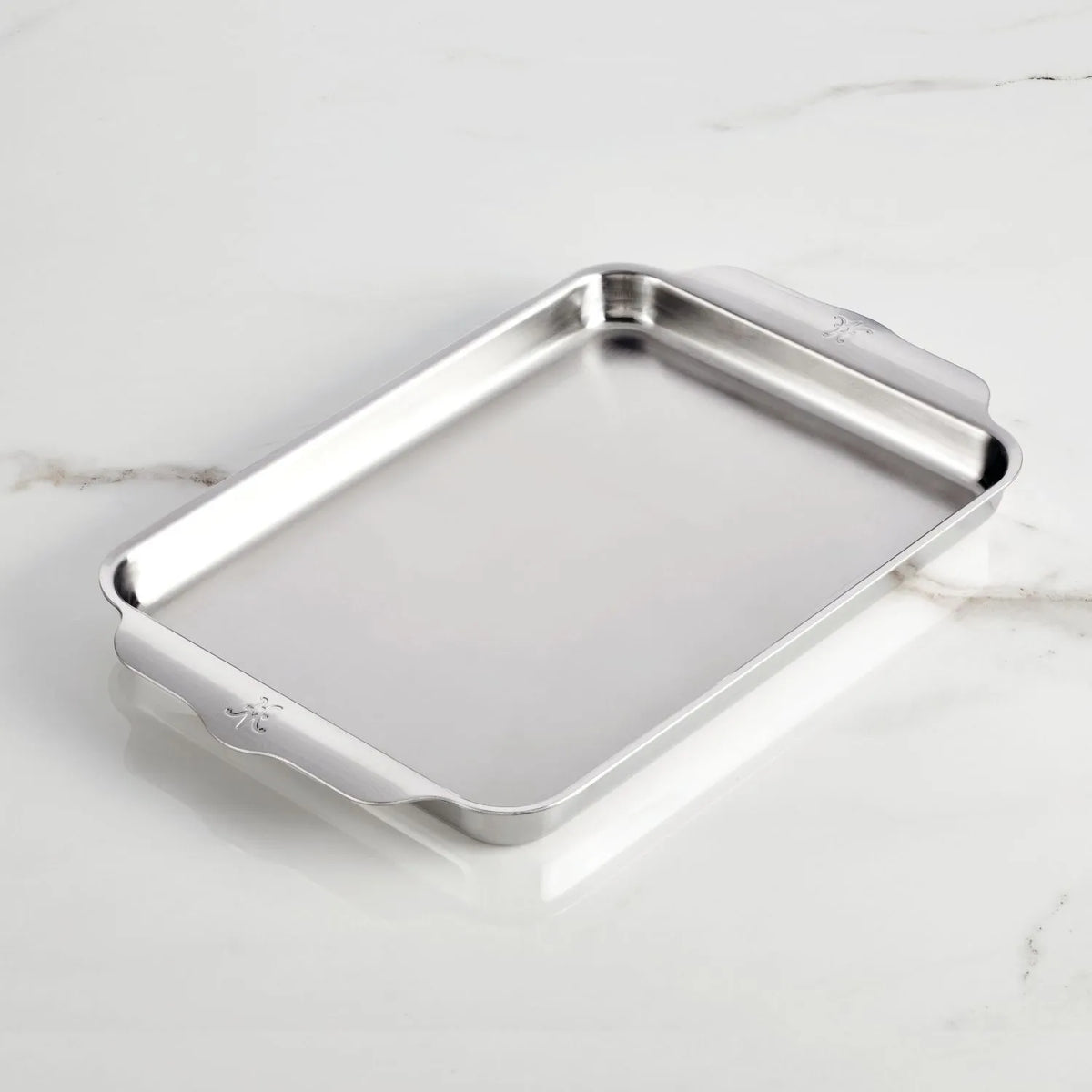 A Hestan OvenBond Tri-Ply Medium Sheet Pan & Rack, 12-in x 15-in, with raised edges and side handles rests on a white marble surface.