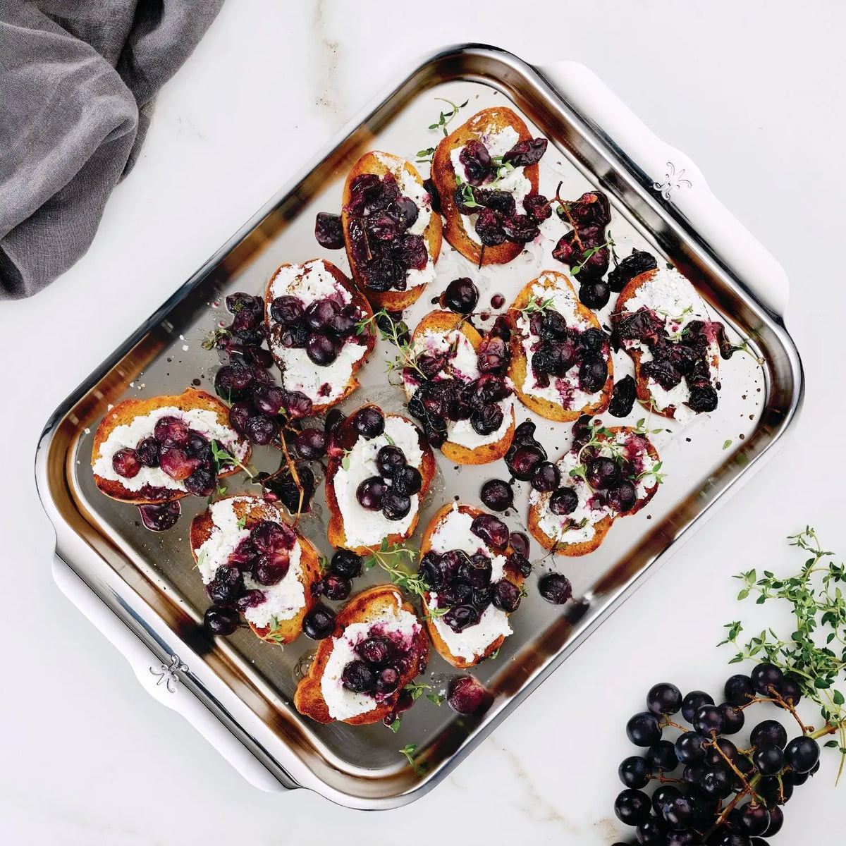 An OvenBond Tri-ply Medium Sheet Pan by Hestan Provisions holds crostini with goat cheese, roasted grapes, and thyme on a marble surface. Nearby are extra grapes and thyme sprigs, with a folded gray napkin to the side.