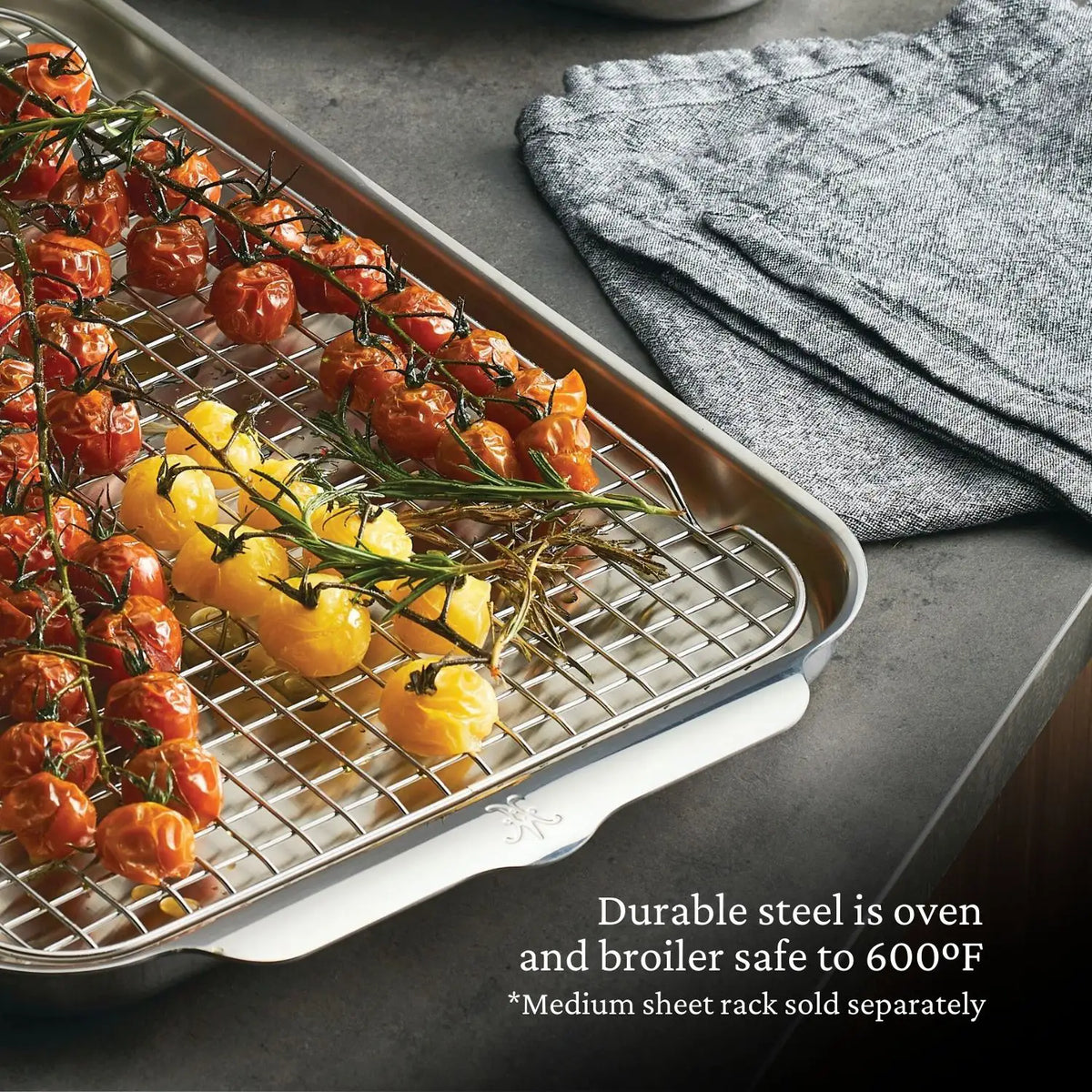 A Hestan Provisions OvenBond Tri-ply Medium Sheet Pan, oven safe to 600ºF, holds roasted cherry tomatoes and yellow tomatoes on a wire rack on the counter. Next to it is a stack of gray kitchen towels, with a note about the wire rack.