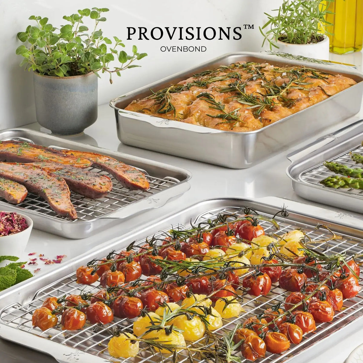 A collection of Hestan Provisions OvenBond Tri-ply Medium Sheet Pans with an array of food on a white surface: one holds roasted tomatoes, another seasoned chicken thighs, a third sliced steak, and the fourth asparagus, with potted herbs visible in the background.