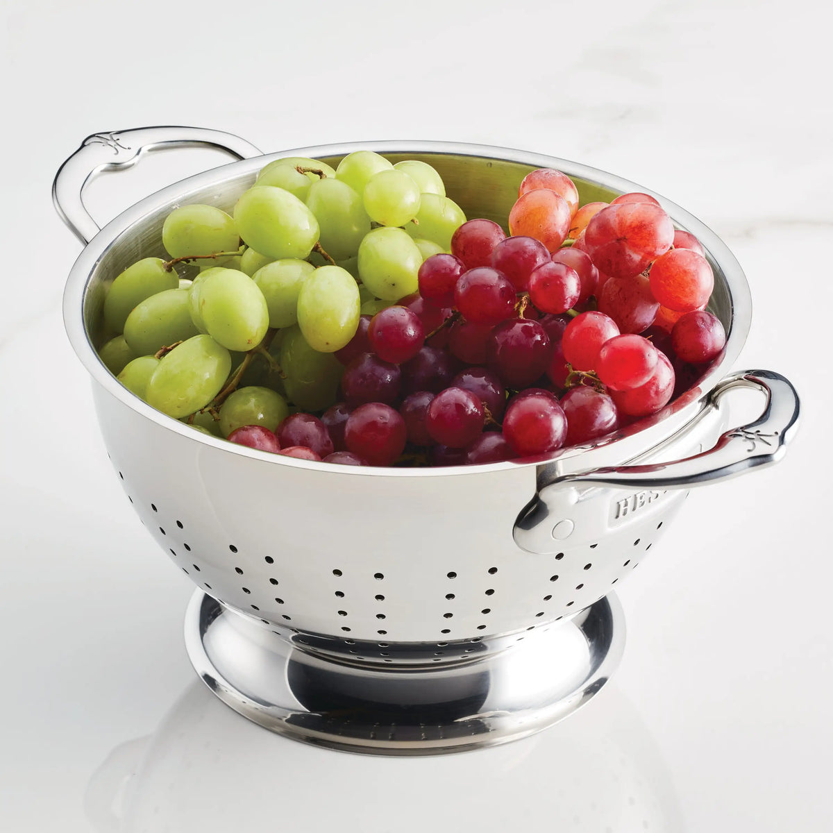 The Hestan Provisions Stainless Steel Colander, shown here with green and red grapes on a white marble surface, is dishwasher safe and provides superior drainage for fresh fruit.