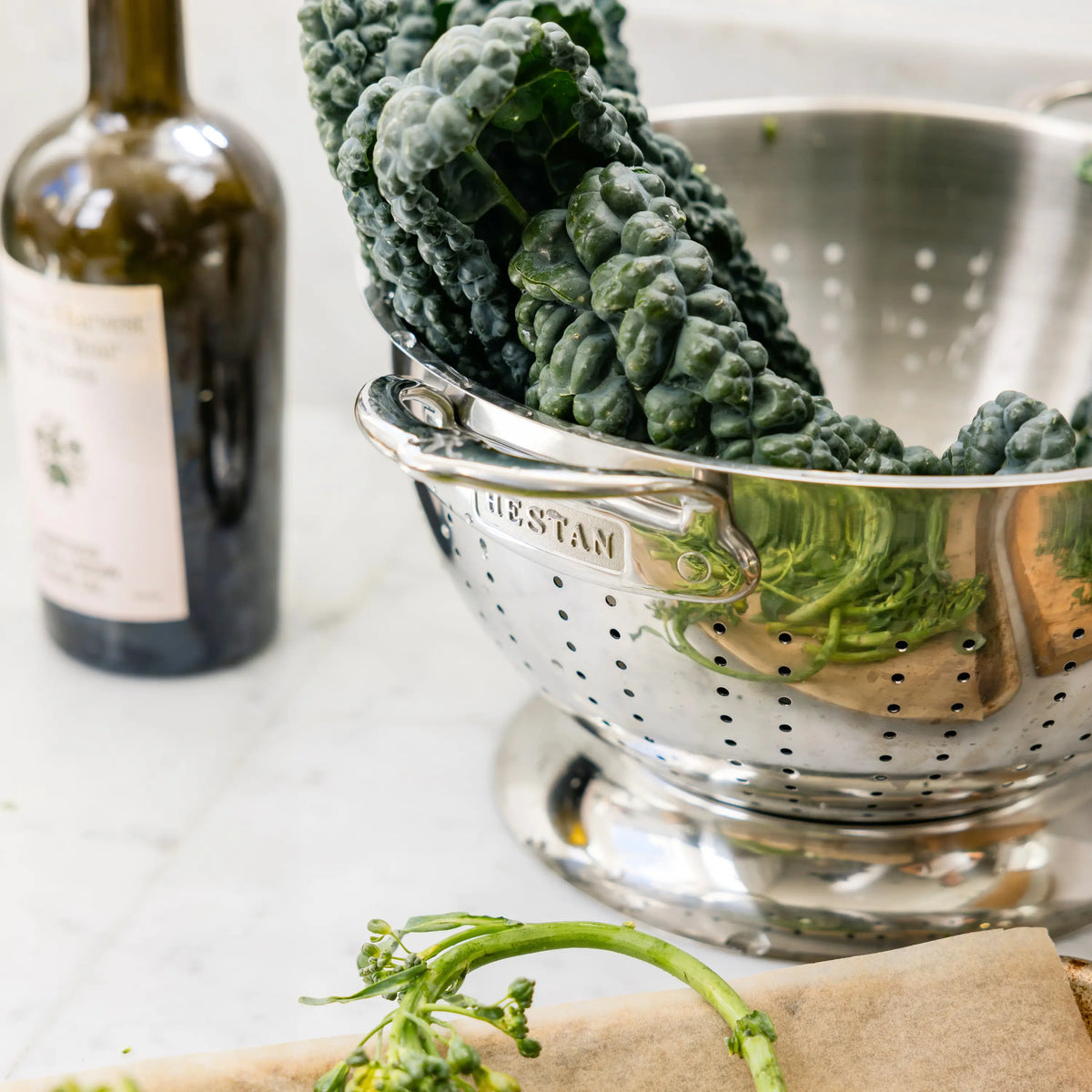 The Hestan Provisions Stainless Steel Colander, filled with dark green kale leaves, sits on a countertop next to olive oil and vegetable stems on brown paper, showcasing its superior drainage.