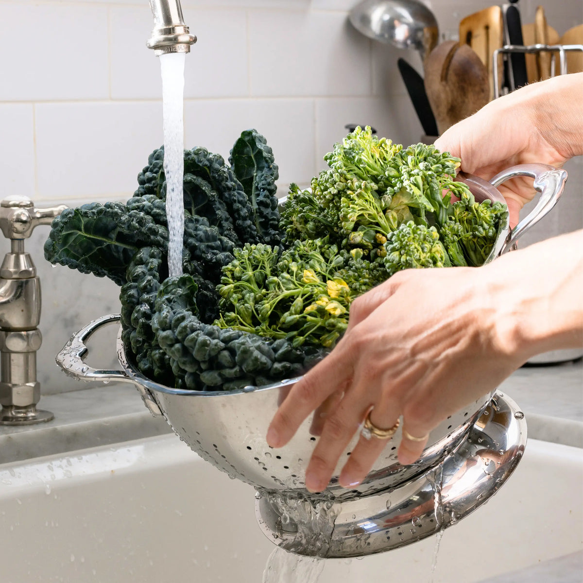 A person rinses leafy greens and broccoli in a Hestan Provisions Stainless Steel Colander with superior drainage under running water at the kitchen sink.