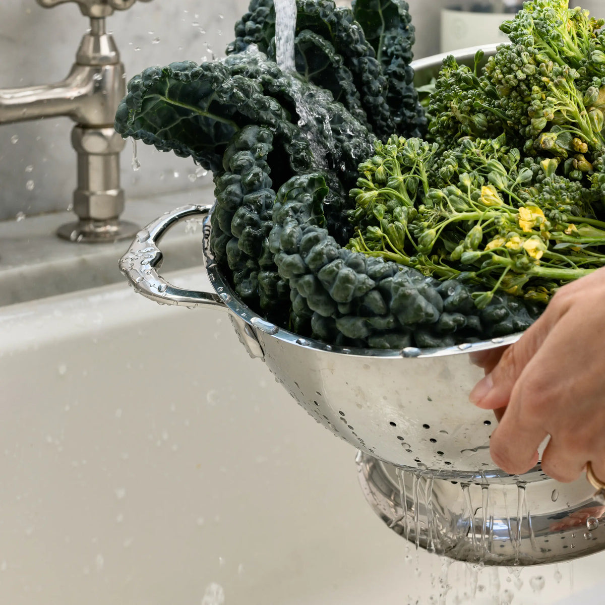 A person rinses leafy greens and broccoli in the Hestan Provisions Stainless Steel Colander under running water, demonstrating its excellent drainage in the kitchen sink.